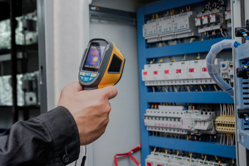 Technician using a handheld thermal imaging camera to inspect an industrial electrical control panel